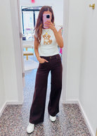 A woman takes a mirror selfie in a hallway, wearing a white tank top with a teddy bear graphic and dark brown wide-leg pants.