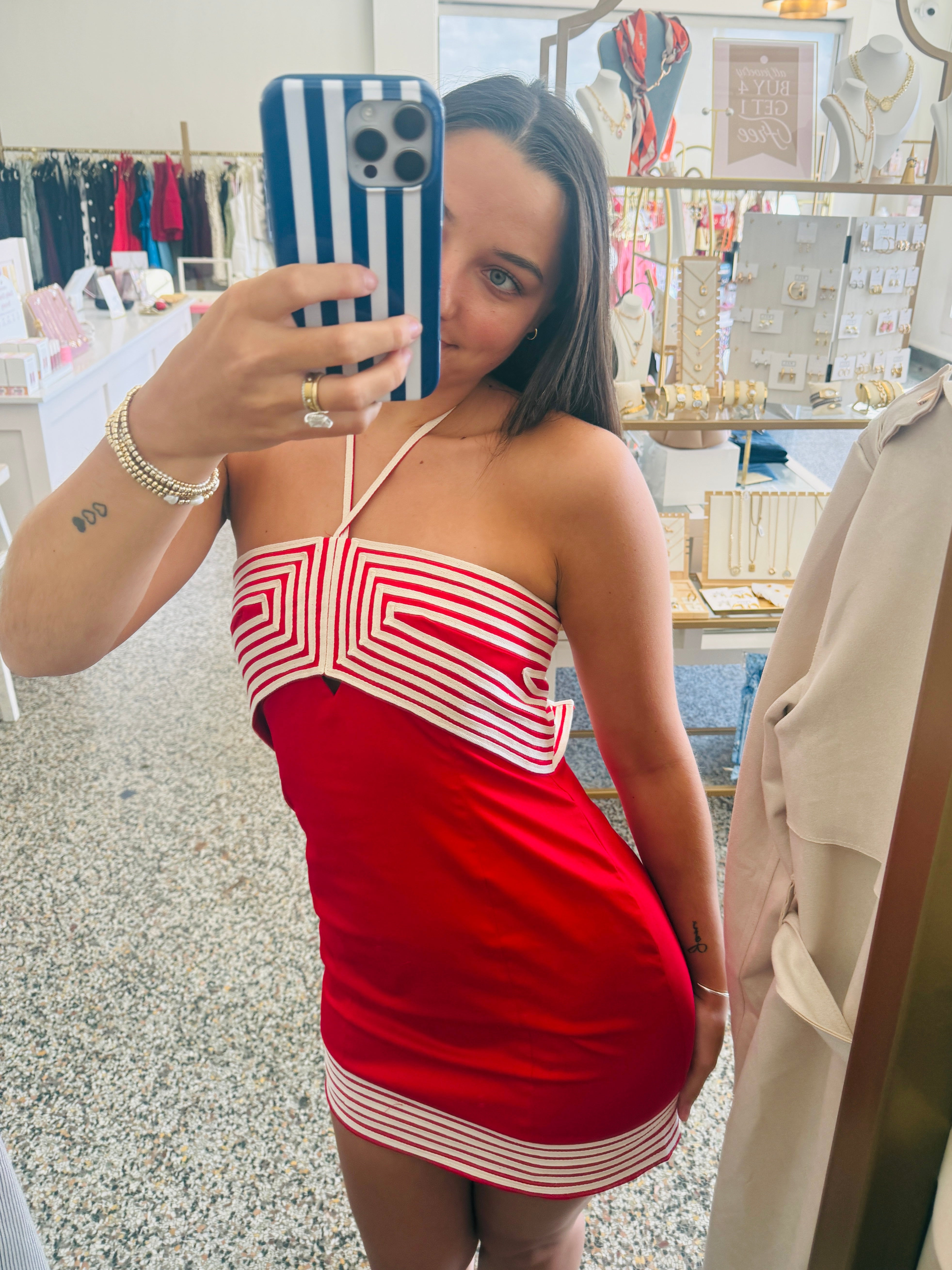 A woman takes a mirror selfie in a boutique while wearing a red mini dress with white geometric stripe patterns. The background shows clothing racks and jewelry displays inside the shop.