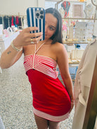 A woman takes a mirror selfie in a boutique while wearing a red mini dress with white geometric stripe patterns. The background shows clothing racks and jewelry displays inside the shop.