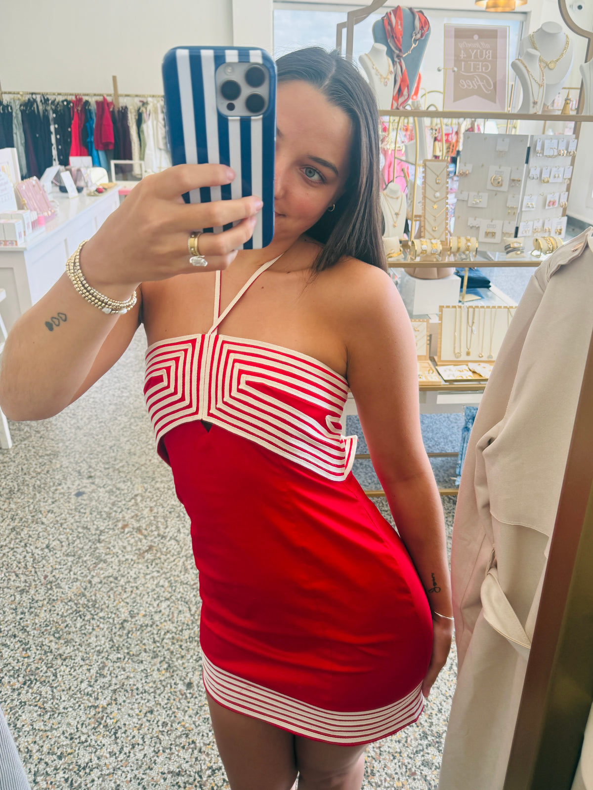 A woman takes a mirror selfie in a boutique while wearing a red mini dress with white geometric stripe patterns. The background shows clothing racks and jewelry displays inside the shop.