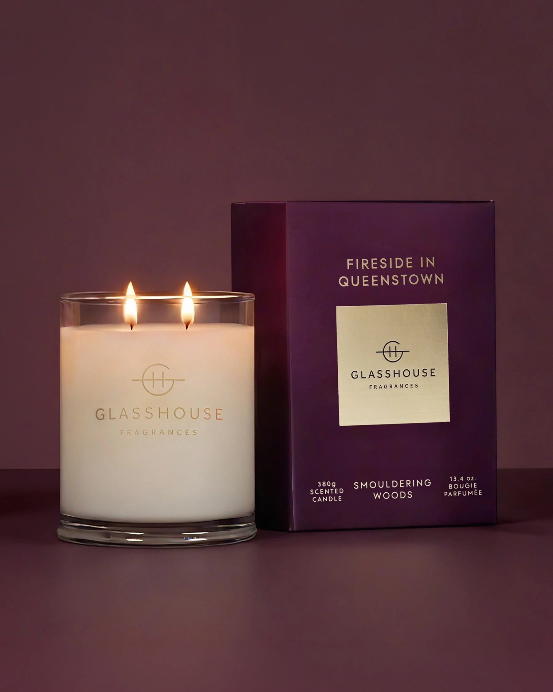 A lit double-wick scented candle in a clear glass jar sits beside its dark purple retail box on a matching purple studio background. The candle and box both show the brand name GLASSHOUSE FRAGRANCES and the scent name FIRESIDE IN QUEENSTOWN.