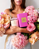 A woman holds a Sea Househouse candle in a glass jar next to its bright pink packaging box, surrounded by large bouquets of pink hydrangeas and peach orchids.