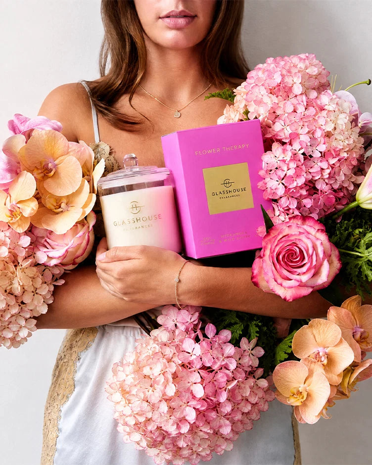 A woman holds a Sea Househouse candle in a glass jar next to its bright pink packaging box, surrounded by large bouquets of pink hydrangeas and peach orchids.