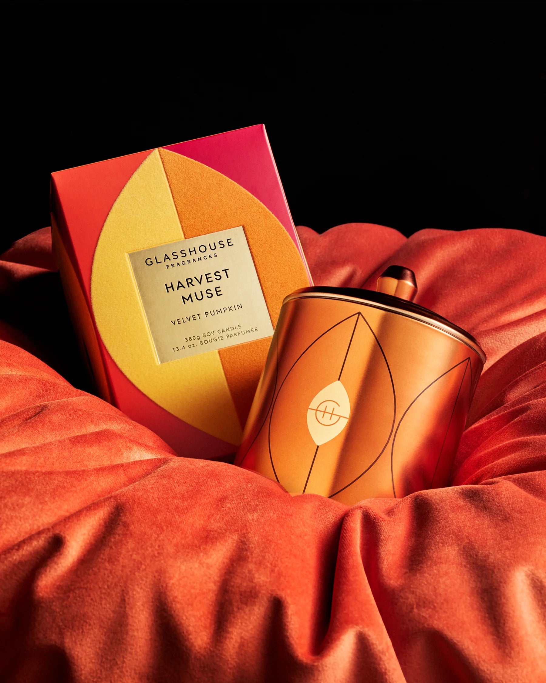 A glass jar candle and its matching box from Glasshouse Aesthetics, Harvest Muse, displayed on a bed of draped rust-orange velvet fabric against a solid black background.