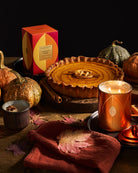 An autumn-themed product still life featuring a Honey Crispin 'Harvest Joy' pumpkin pie, a lit copper candle, a small candle in a mug, and a decorative box, styled with pumpkins, fall leaves, and a rust-colored cloth on a dark wooden table.