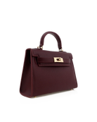 A structured mini handbag in deep burgundy textured leather with gold-tone hardware, featuring a top handle and front flap closure, shown on a plain white background.