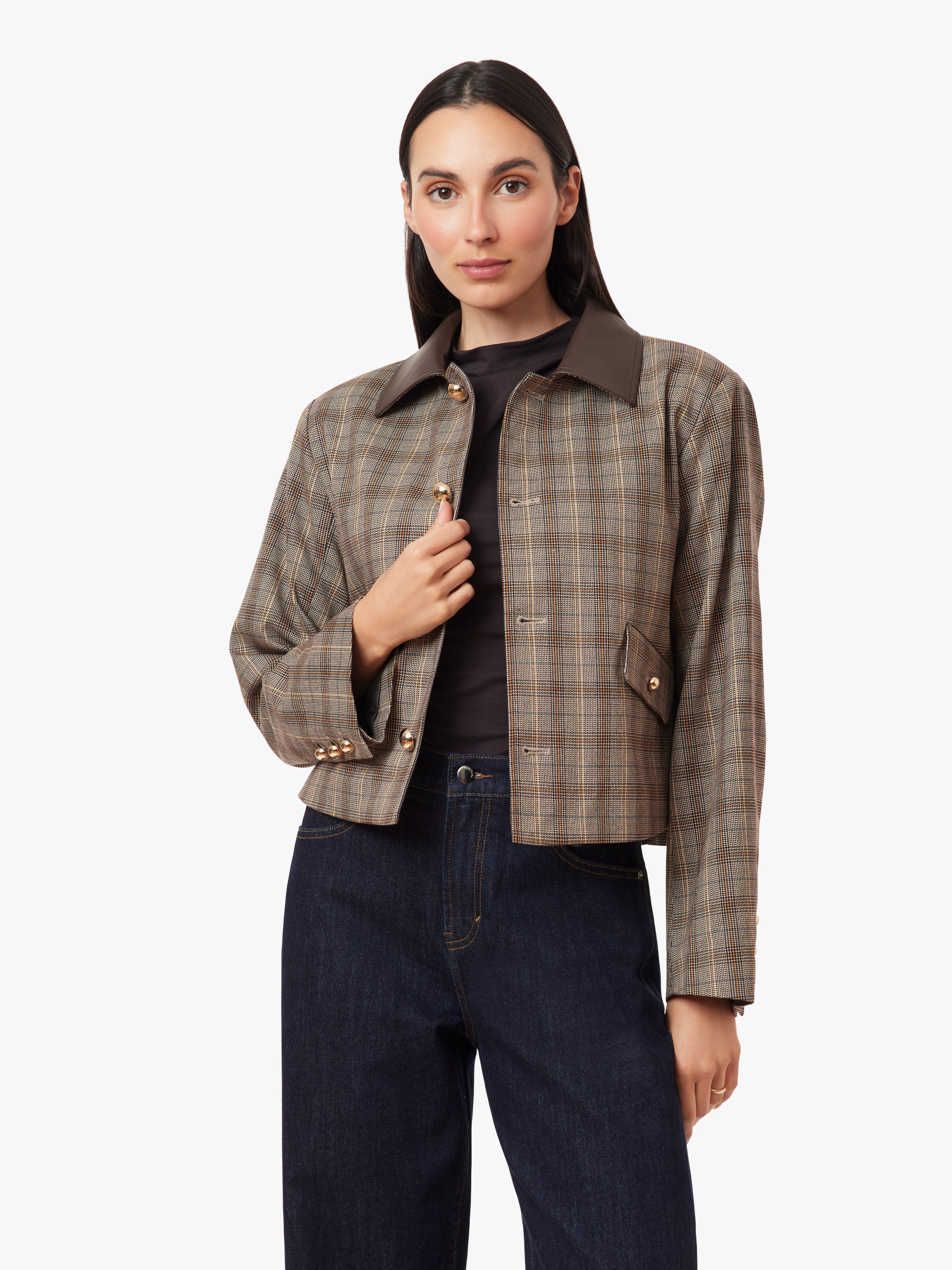 A woman modeling a cropped plaid jacket with a dark brown collar, worn over a black top and dark blue jeans, against a plain white studio background.