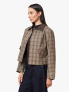 A woman wearing a cropped plaid jacket with a dark brown collar, styled with a black top and dark blue jeans, photographed against a light gray studio background.