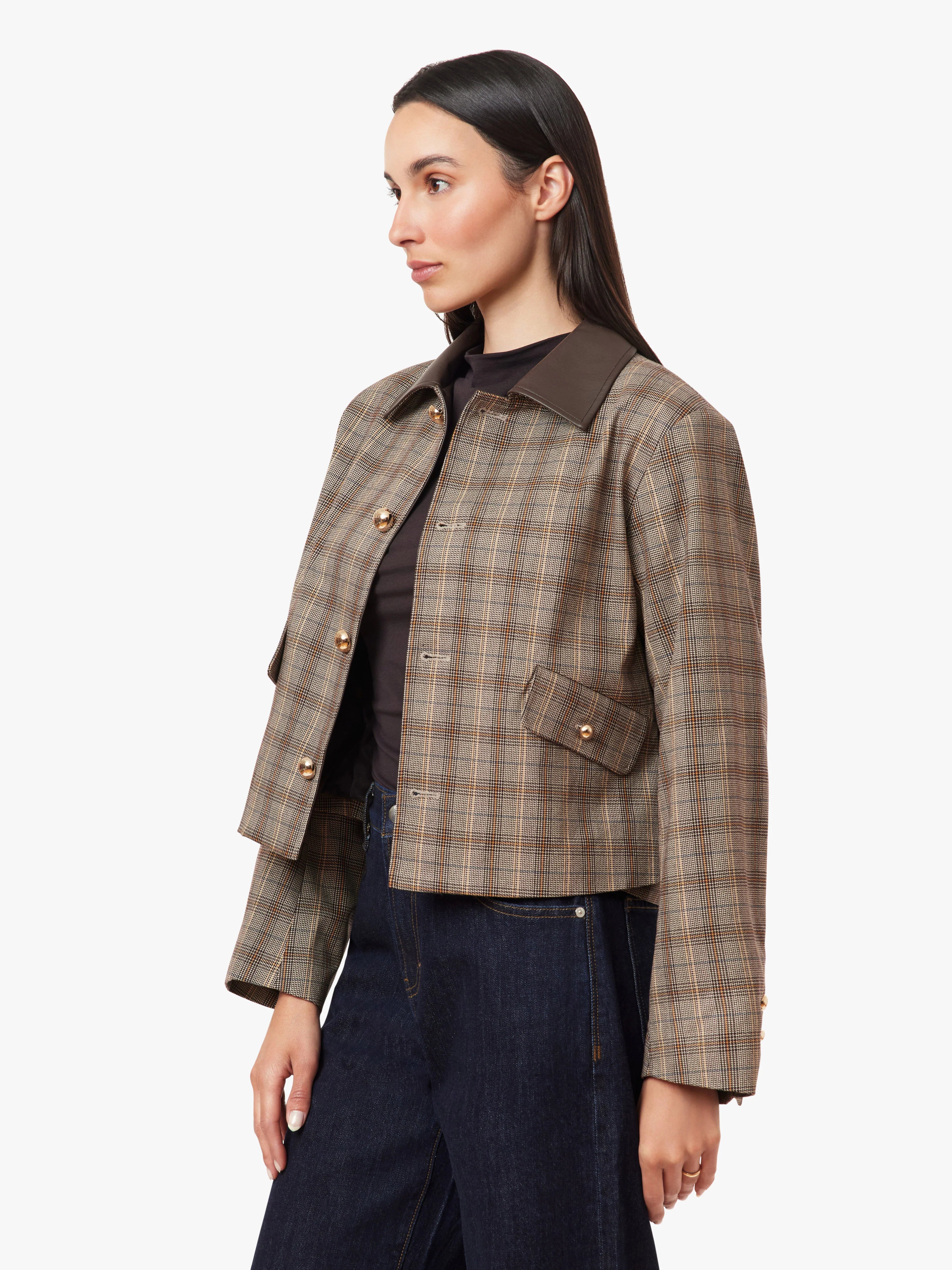 A woman wearing a cropped plaid jacket with a dark brown collar, styled with a black top and dark blue jeans, photographed against a light gray studio background.