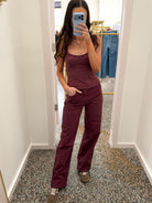 A woman takes a mirror selfie wearing a matching two-piece set consisting of a sleeveless scoop-neck camisole tank top and wide-leg trousers, both in a deep burgundy color. She is standing in a fitting room with a speckled floor and a clothing rack visible in the background.