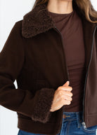 A close-up of a person wearing a dark brown jacket with a fuzzy shearling-style collar and matching cuffs, layered over a brown top and blue jeans.