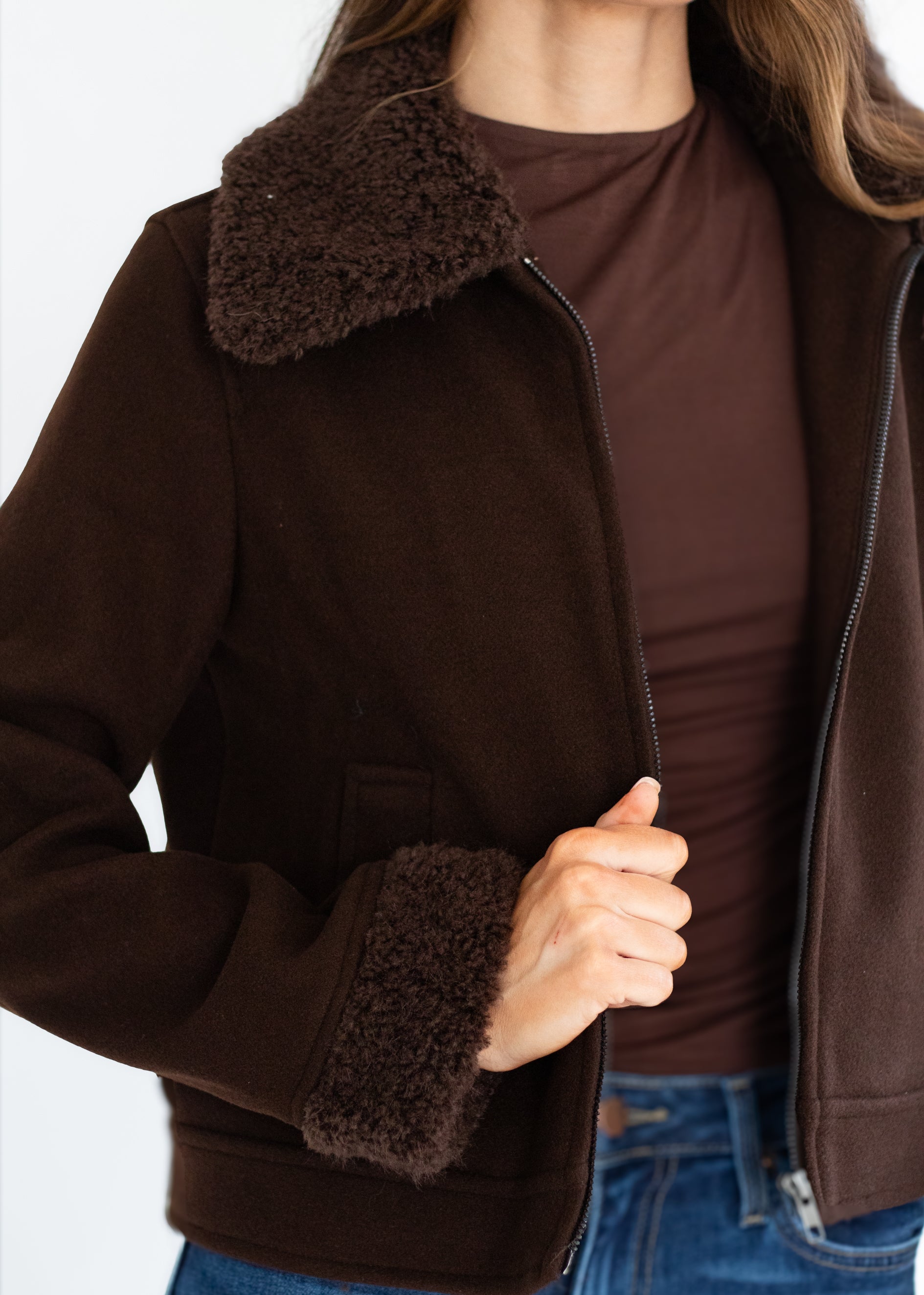 A close-up of a person wearing a dark brown jacket with a fuzzy shearling-style collar and matching cuffs, layered over a brown top and blue jeans.