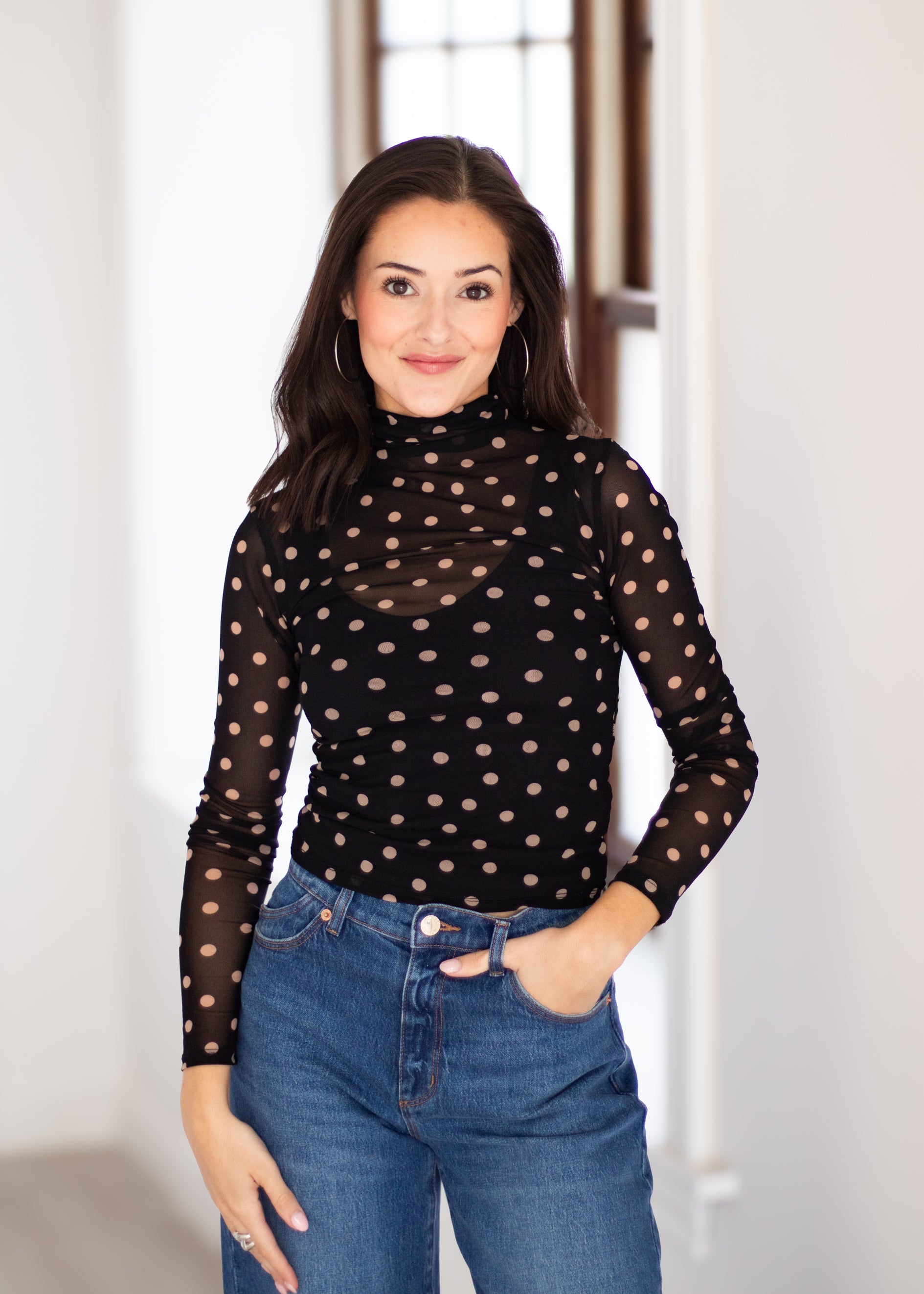 A woman wearing a black sheer long-sleeve mock neck top with beige polka dots, paired with blue jeans, standing in a brightly lit indoor setting.