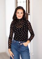 A woman wearing a black sheer long-sleeve mock neck top with beige polka dots, paired with blue jeans, standing in a brightly lit indoor setting.