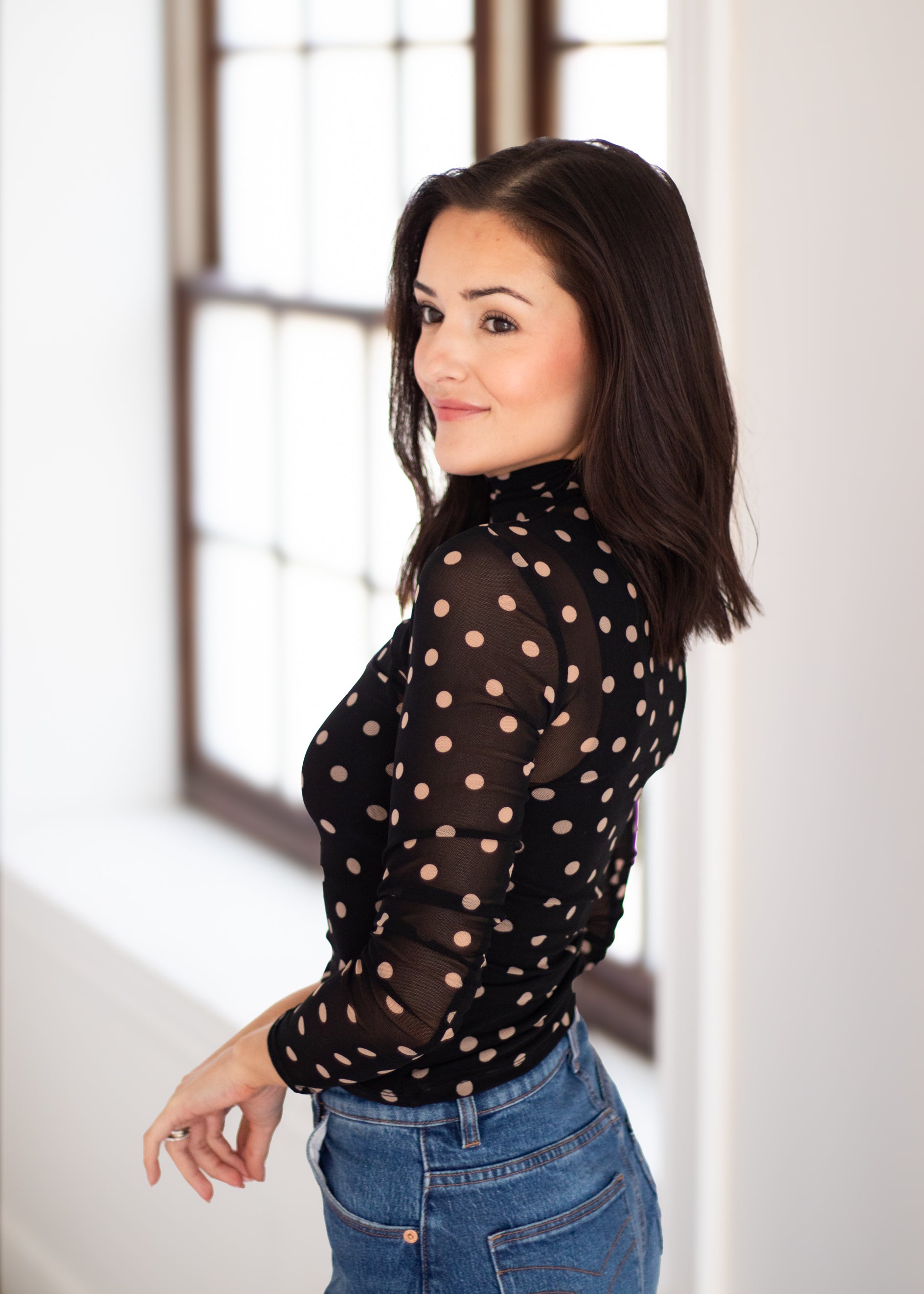 A woman with dark hair is shown from the back and side, wearing a sheer black long-sleeve top with beige polka dots and blue jeans, standing in a brightly lit room with a window.