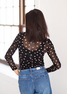 A woman viewed from behind wearing a sheer black long-sleeve top with beige polka dots and blue jeans, standing in a bright indoor setting with a window in the background.