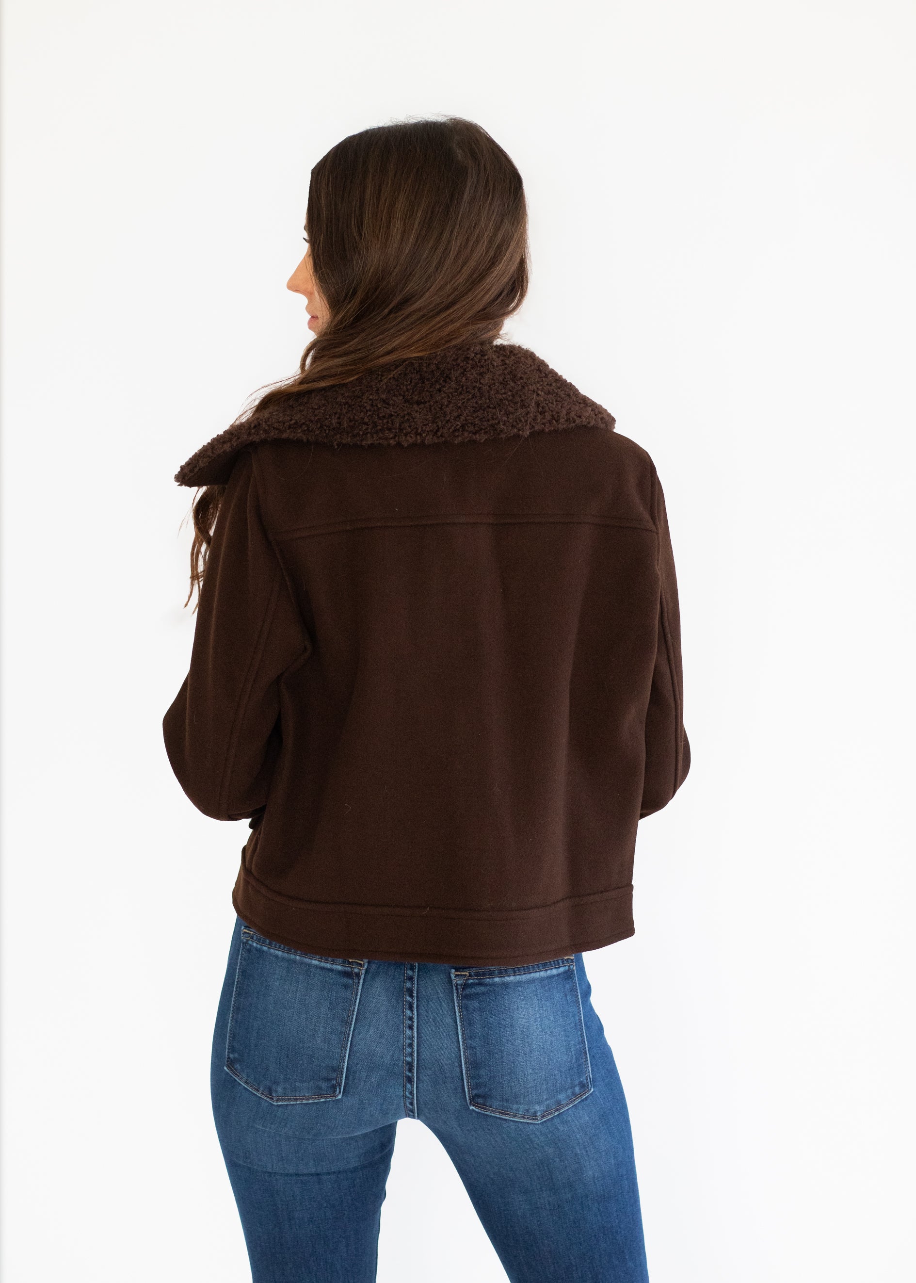 A woman shown from the back wearing a dark brown cropped jacket with a fuzzy brown shearling-style collar and blue jeans, against a plain white studio background.
