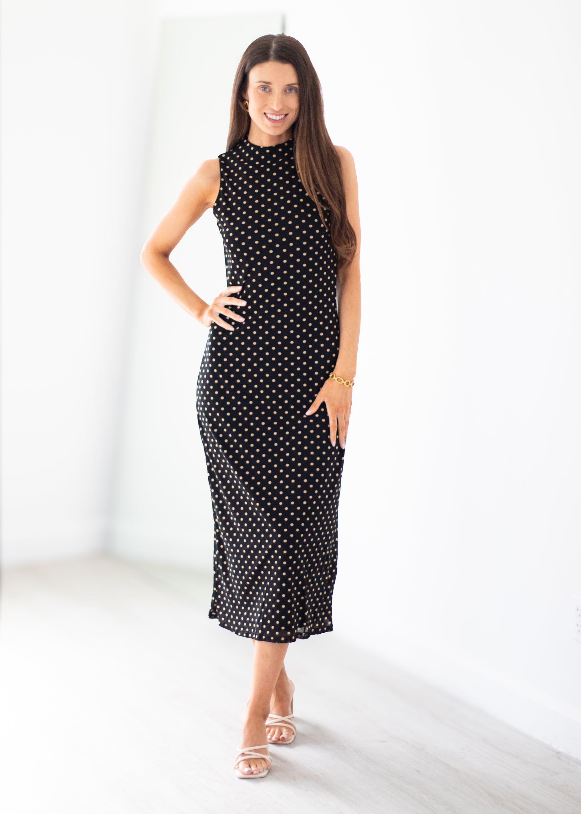 A woman modeling a sleeveless black midi dress with a white polka dot pattern, standing in a bright, minimalist white room.