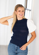 A smiling blonde woman wearing a navy blue open-knit sleeveless sweater vest over a white short-sleeve blouse and blue jeans, standing in a bright indoor setting.