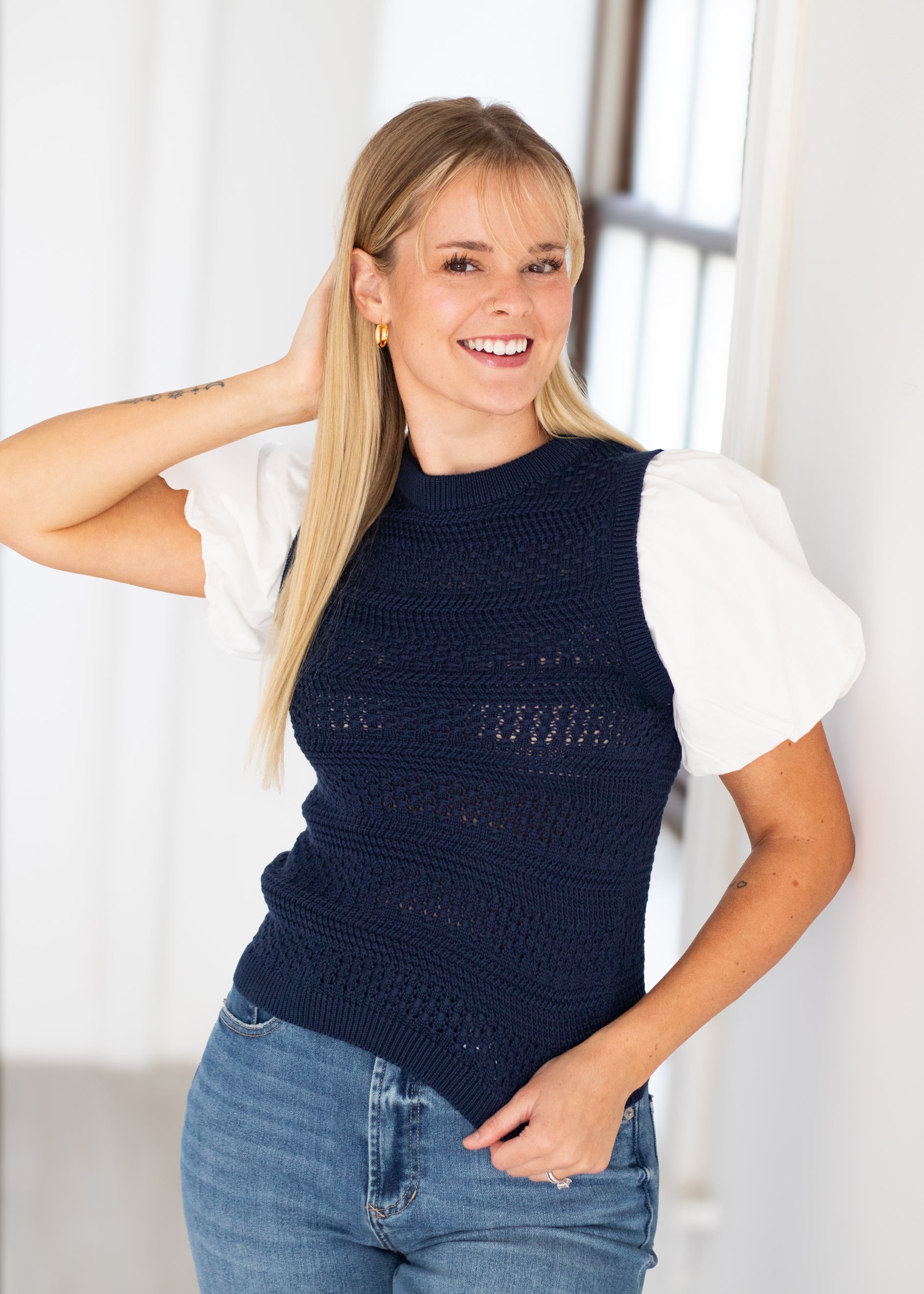 A smiling blonde woman wearing a navy blue open-knit sleeveless sweater vest over a white short-sleeve blouse and blue jeans, standing in a bright indoor setting.