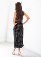 A woman stands with her back to the camera wearing a sleeveless black midi dress with a white polka dot pattern and a lace-up open back, paired with white block-heel sandals in a bright white room.