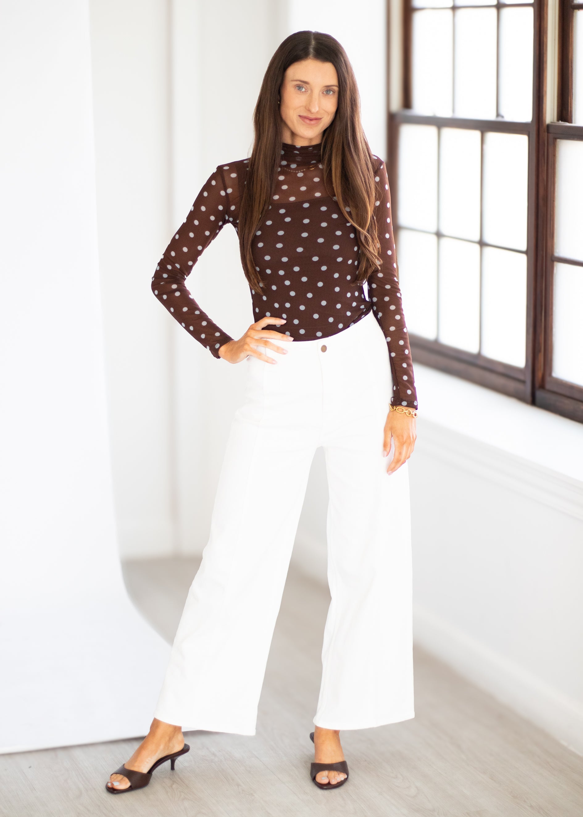 A woman modeling a brown long-sleeve sheer mesh top with small white polka dots, paired with white wide-leg pants and black heeled sandals, standing in a bright room with a large window.
