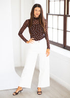 A woman modeling a brown long-sleeve sheer mesh top with small white polka dots, paired with white wide-leg pants and black heeled sandals, standing in a bright room with a large window.