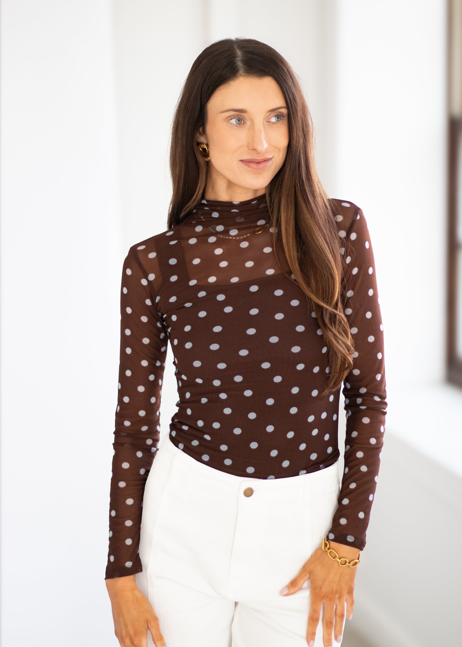 A woman wearing a brown mock-neck long-sleeve top with a white polka-dot pattern, paired with white pants, standing in a brightly lit indoor setting.