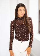 A woman wearing a brown mock-neck long-sleeve top with a white polka-dot pattern, paired with white pants, standing in a brightly lit indoor setting.