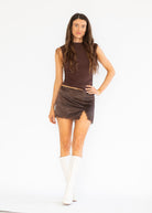 A woman modeling a dark brown sleeveless mock-neck top and a matching dark brown ruched mini skirt with raw-edge detailing, paired with white knee-high boots, against a plain white studio background.