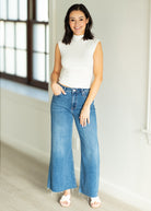 Woman wearing a white sleeveless top and blue jeans standing in a well-lit room.