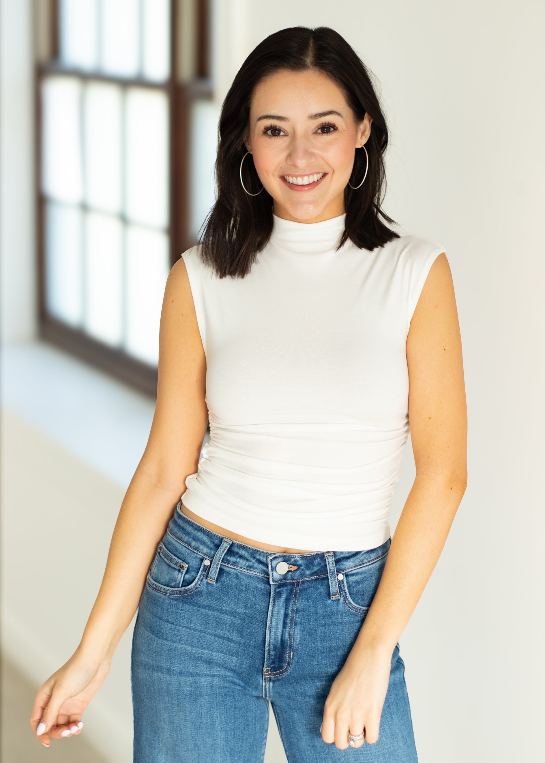 Woman wearing a white sleeveless top and blue jeans indoors.