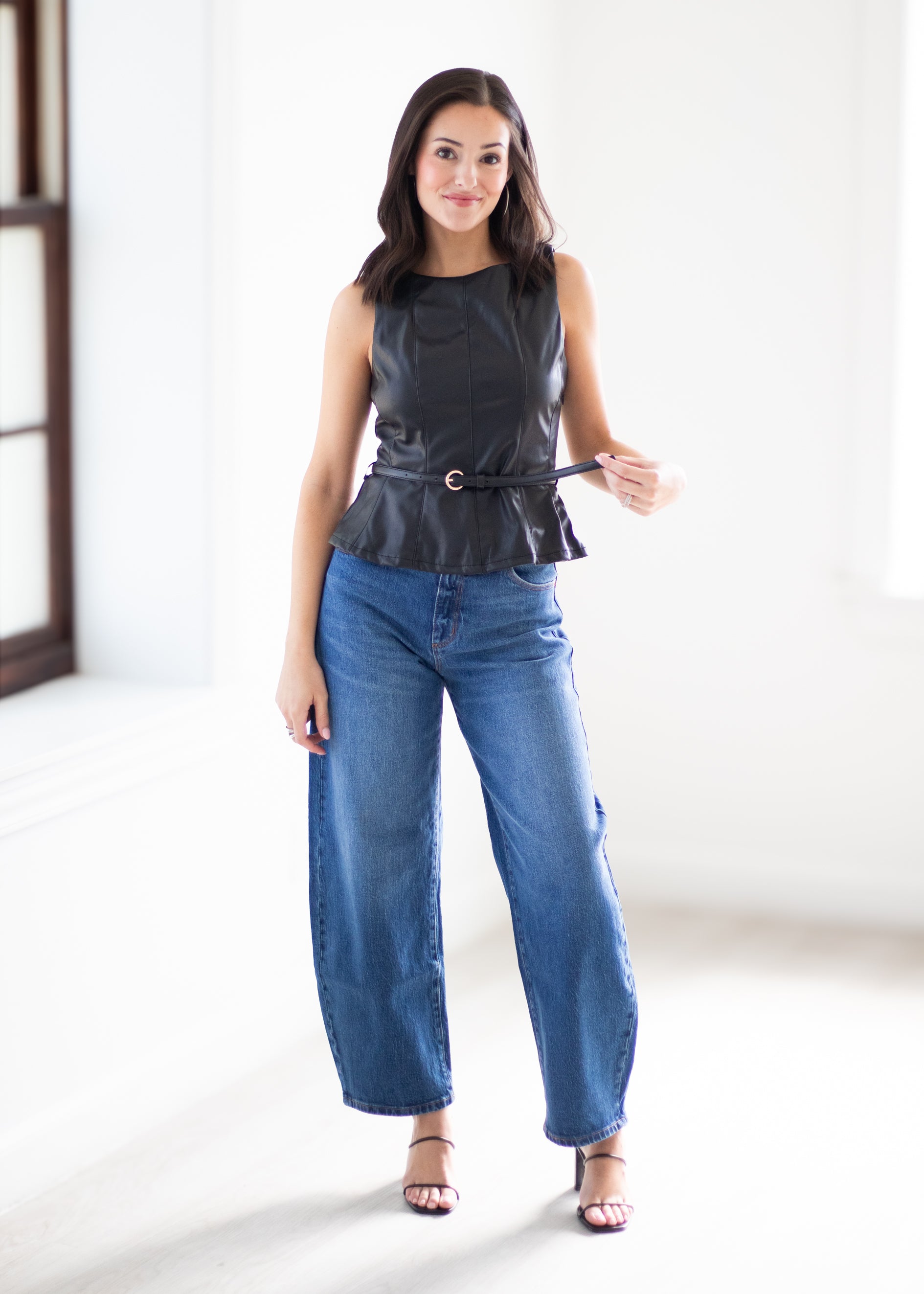 A woman modeling a sleeveless black faux-leather peplum top with a matching belt and blue wide-leg jeans in a bright, minimalist room.