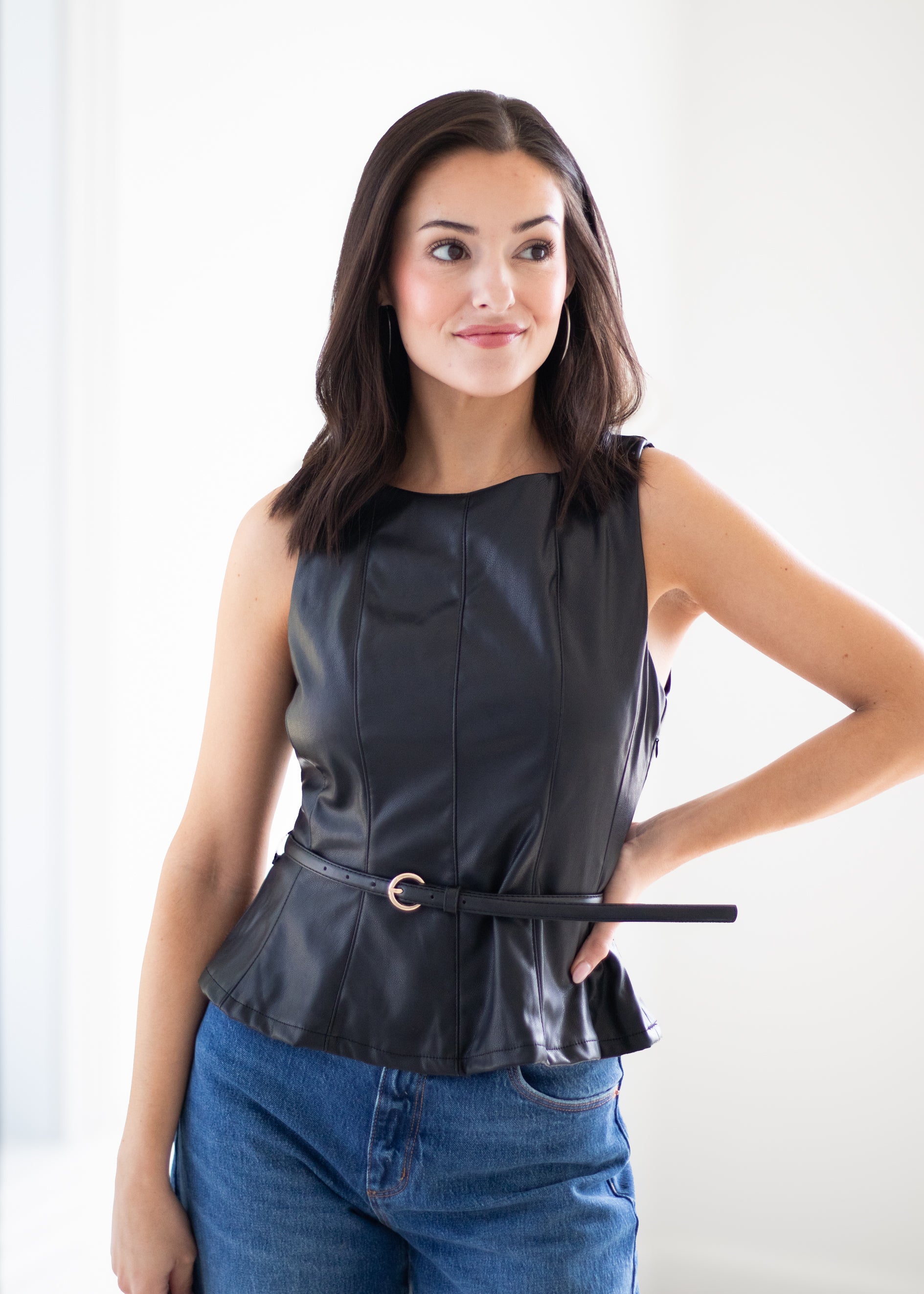 A woman modeling a black faux-leather sleeveless peplum top with a matching belt, paired with blue jeans, against a bright white background.