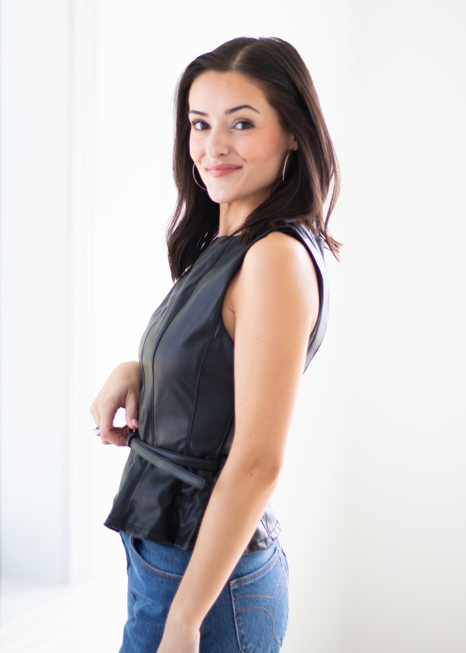 A woman wearing a sleeveless black faux-leather peplum top and blue jeans, posing in a bright white room.