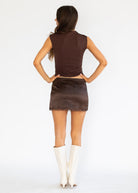 A woman shown from the back wearing a dark brown sleeveless top and matching mini skirt with a scalloped hem, paired with white knee-high boots, against a plain white studio background.