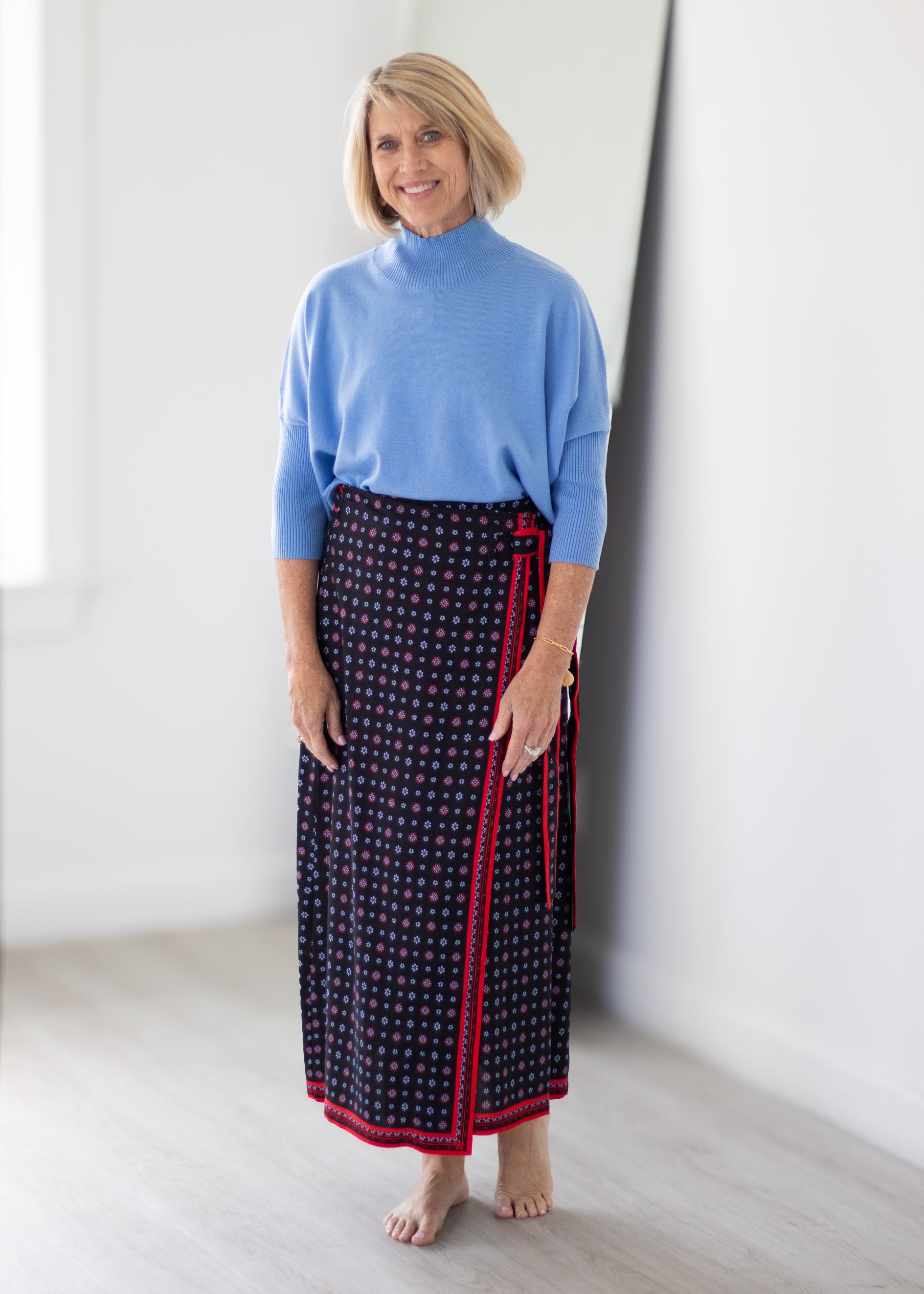 A woman stands barefoot in a bright white room, wearing a light blue mock-neck knit top and a dark navy wrap skirt with a red and white pattern and red trim.