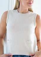 A woman wearing a sleeveless ribbed knit sweater vest in a light cream or off-white color, featuring two front patch pockets and decorative side slit details.