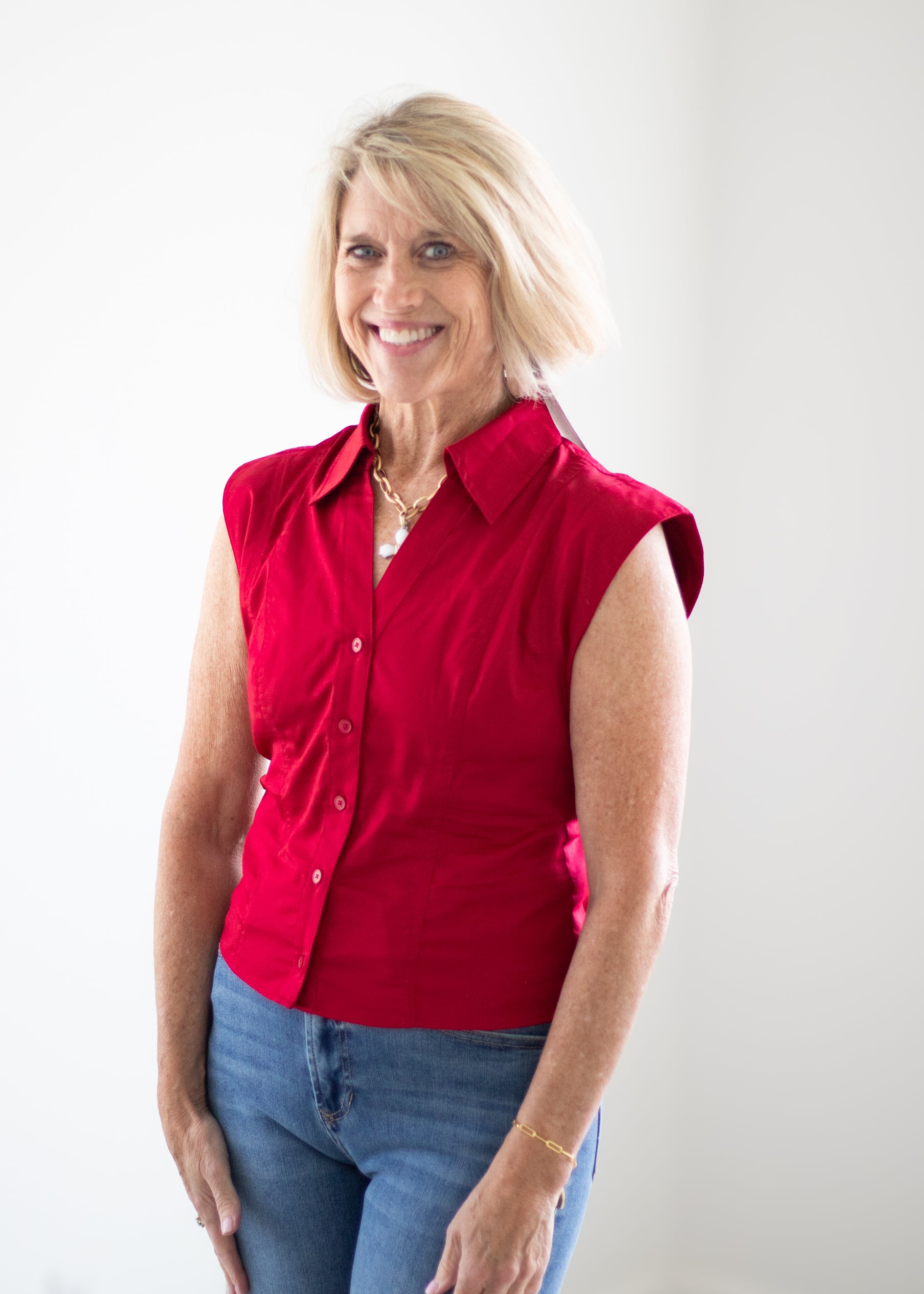 A smiling blonde woman wearing a sleeveless red button-up shirt and blue jeans, standing against a plain white background.