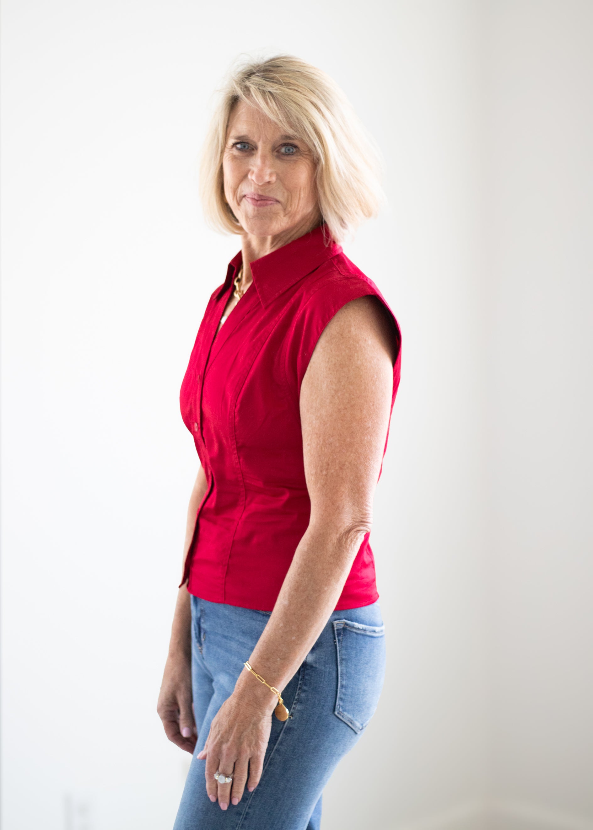 A woman with blonde hair wearing a sleeveless red button-up blouse and blue jeans, standing against a plain white wall.