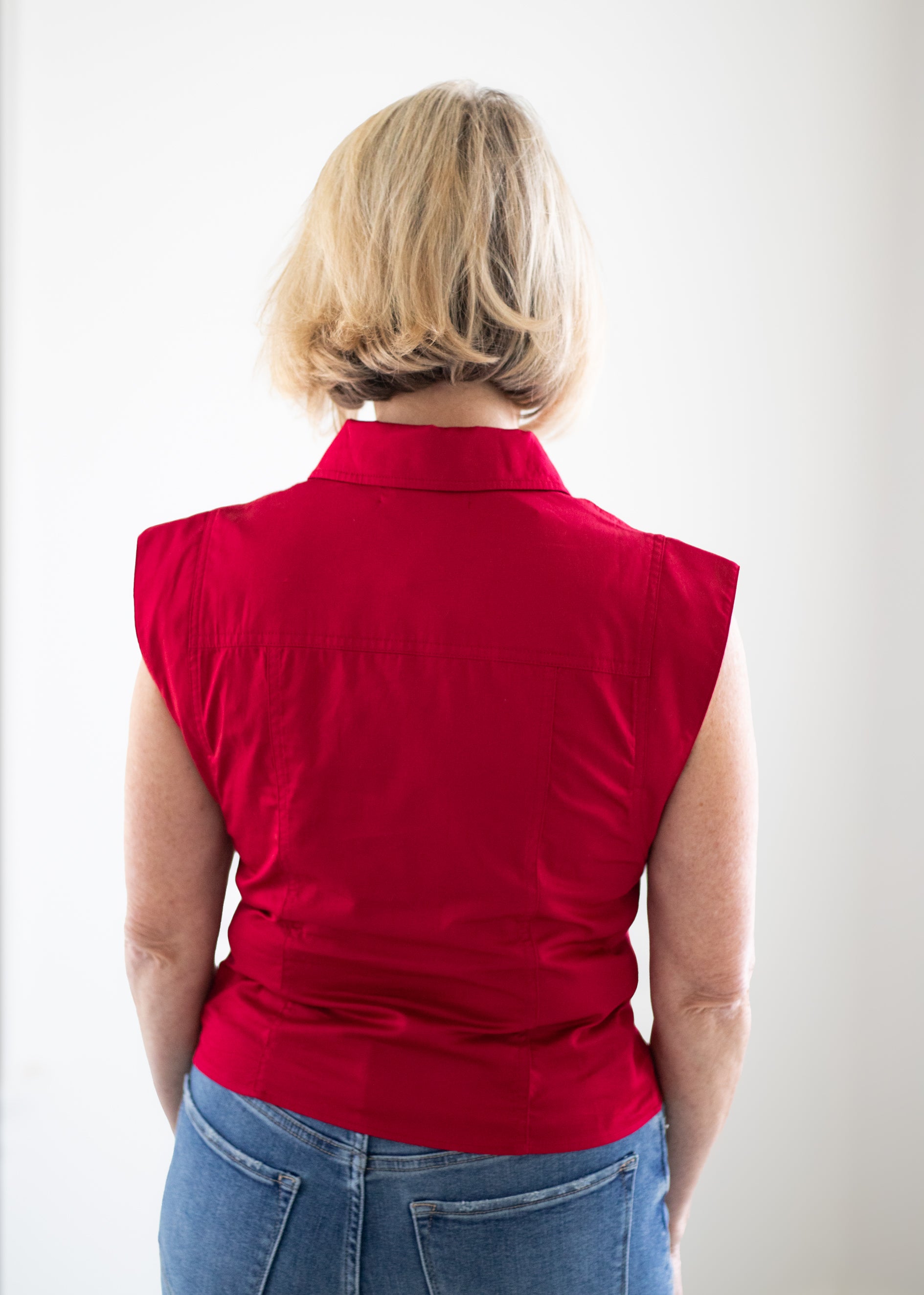 A woman viewed from behind wearing a sleeveless red collared top and blue jeans against a plain white background.