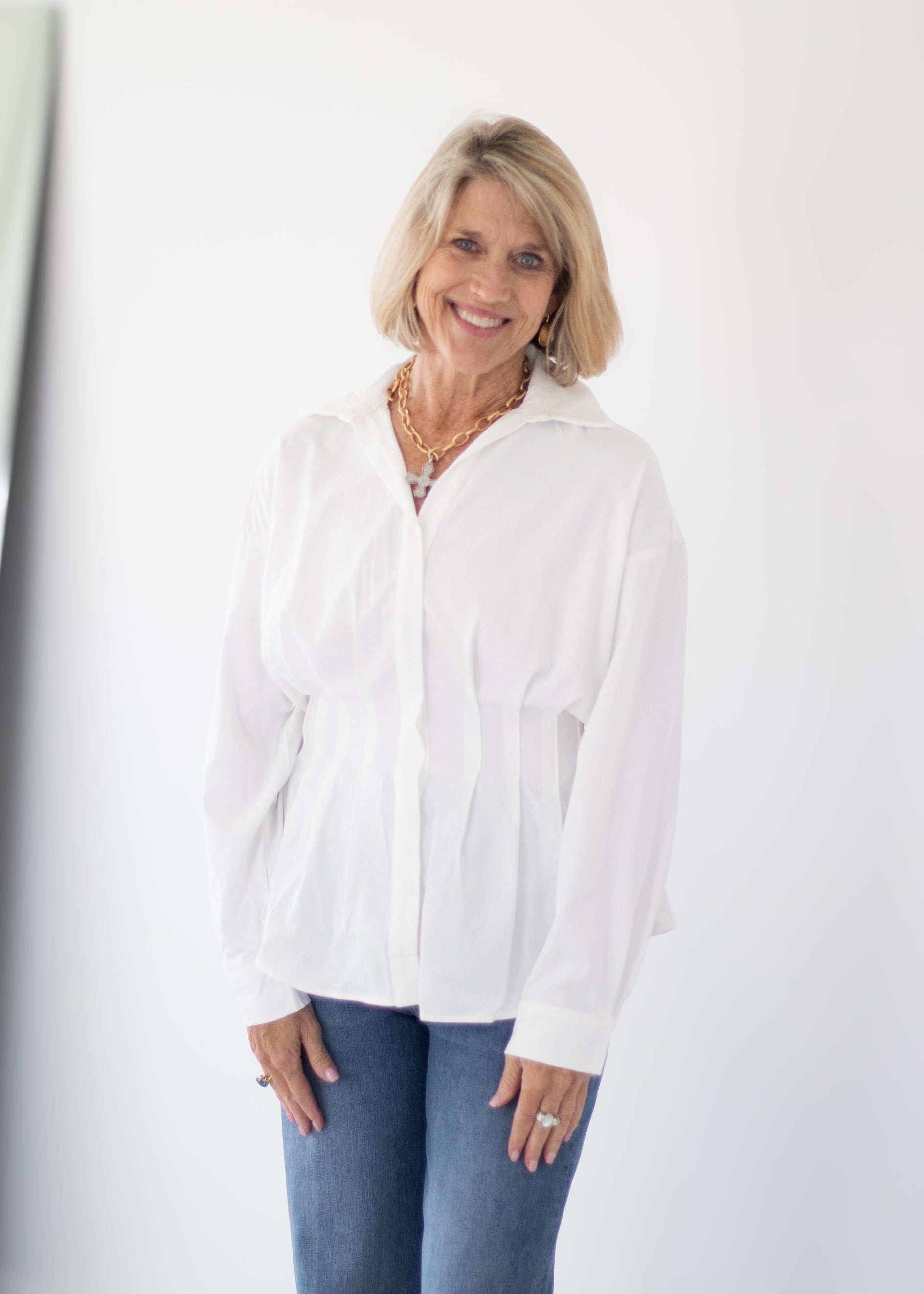 A smiling blonde woman wearing a white long-sleeve button-up shirt and blue jeans, standing against a plain white wall.