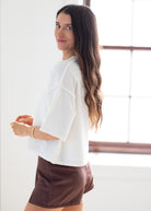 A woman with long brown hair is shown from the side and back, wearing a white short-sleeve cropped top and brown shorts in a bright indoor setting with a window.