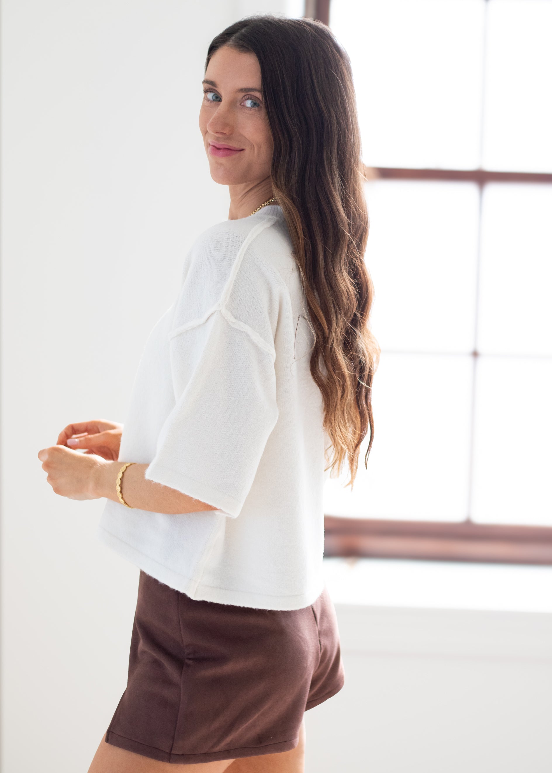 A woman with long brown hair is shown from the side and back, wearing a white short-sleeve cropped top and brown shorts in a bright indoor setting with a window.