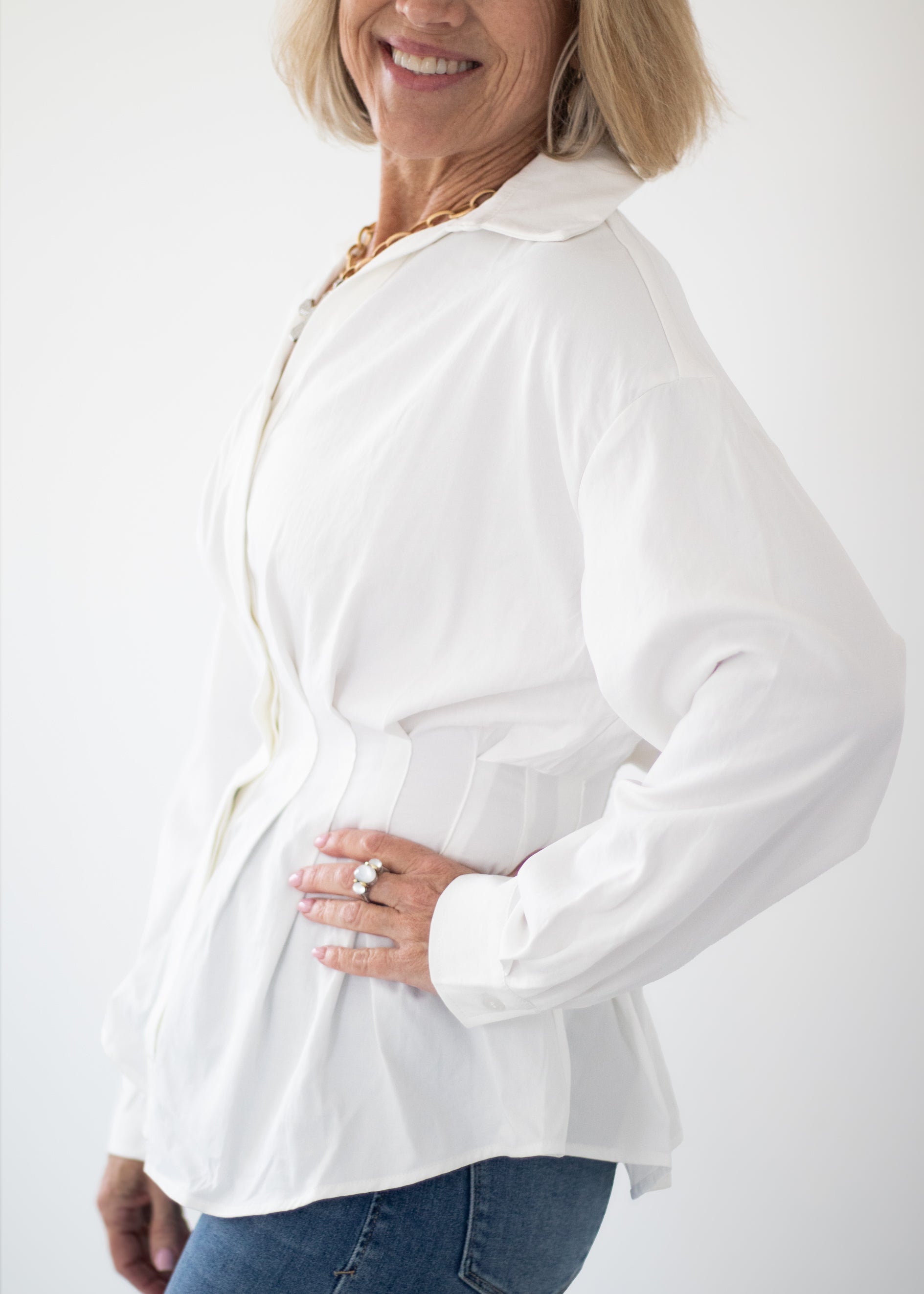 A woman wearing a white long-sleeve collared blouse with a button-front closure and side pockets, paired with blue jeans, shown against a plain light background.