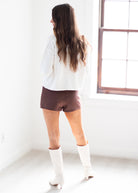 A woman stands with her back to the camera in a bright white room, wearing a white long-sleeve top, brown shorts, and white knee-high boots.