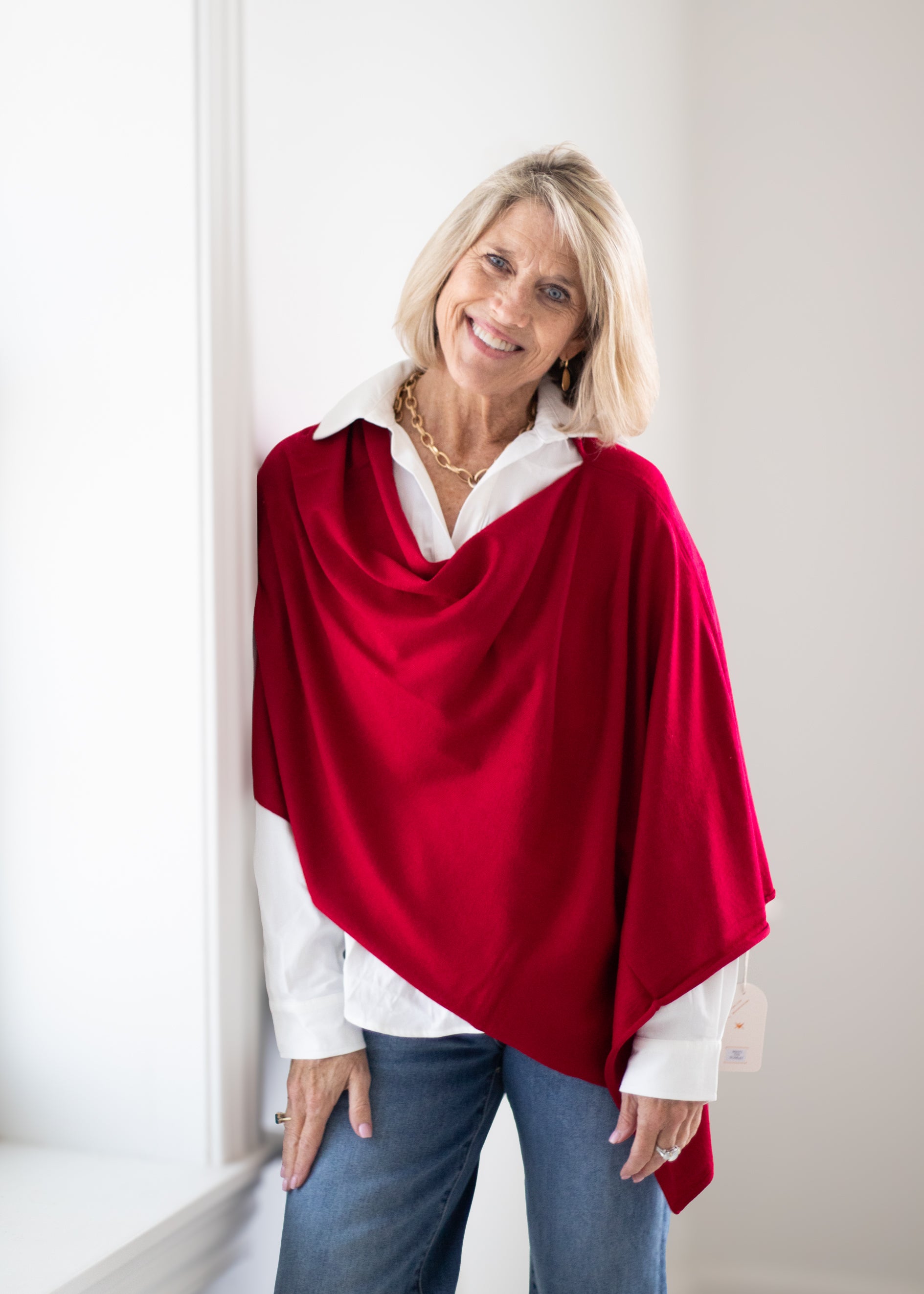 A smiling blonde woman wearing a bright red cowl-neck wrap poncho over a white collared shirt and blue jeans, standing in a brightly lit indoor setting.