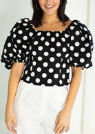 Woman wearing a black polka dot blouse with white pants against a light background