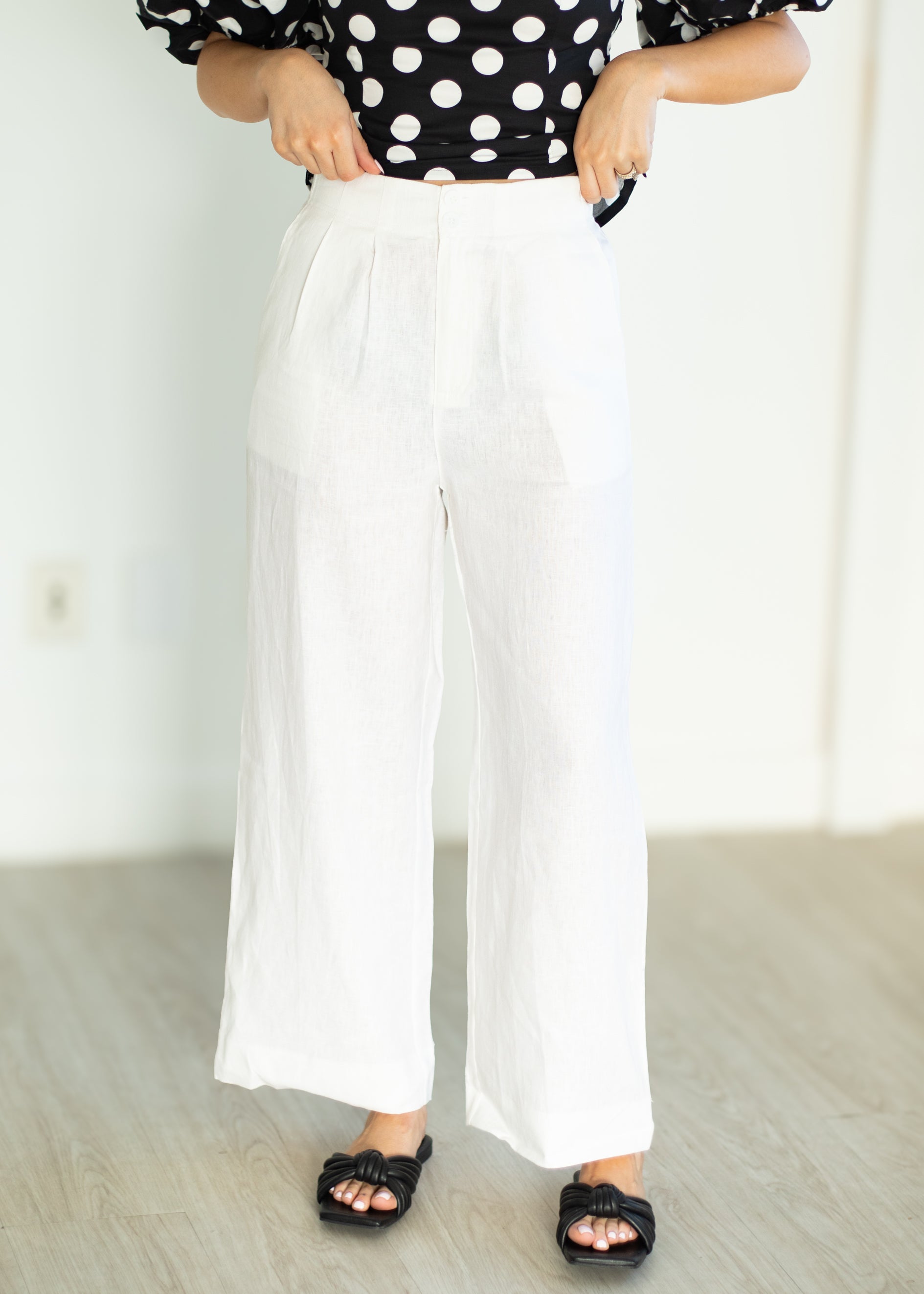 Person wearing a black and white polka dot shirt with white wide-leg pants in a minimal indoor setting.