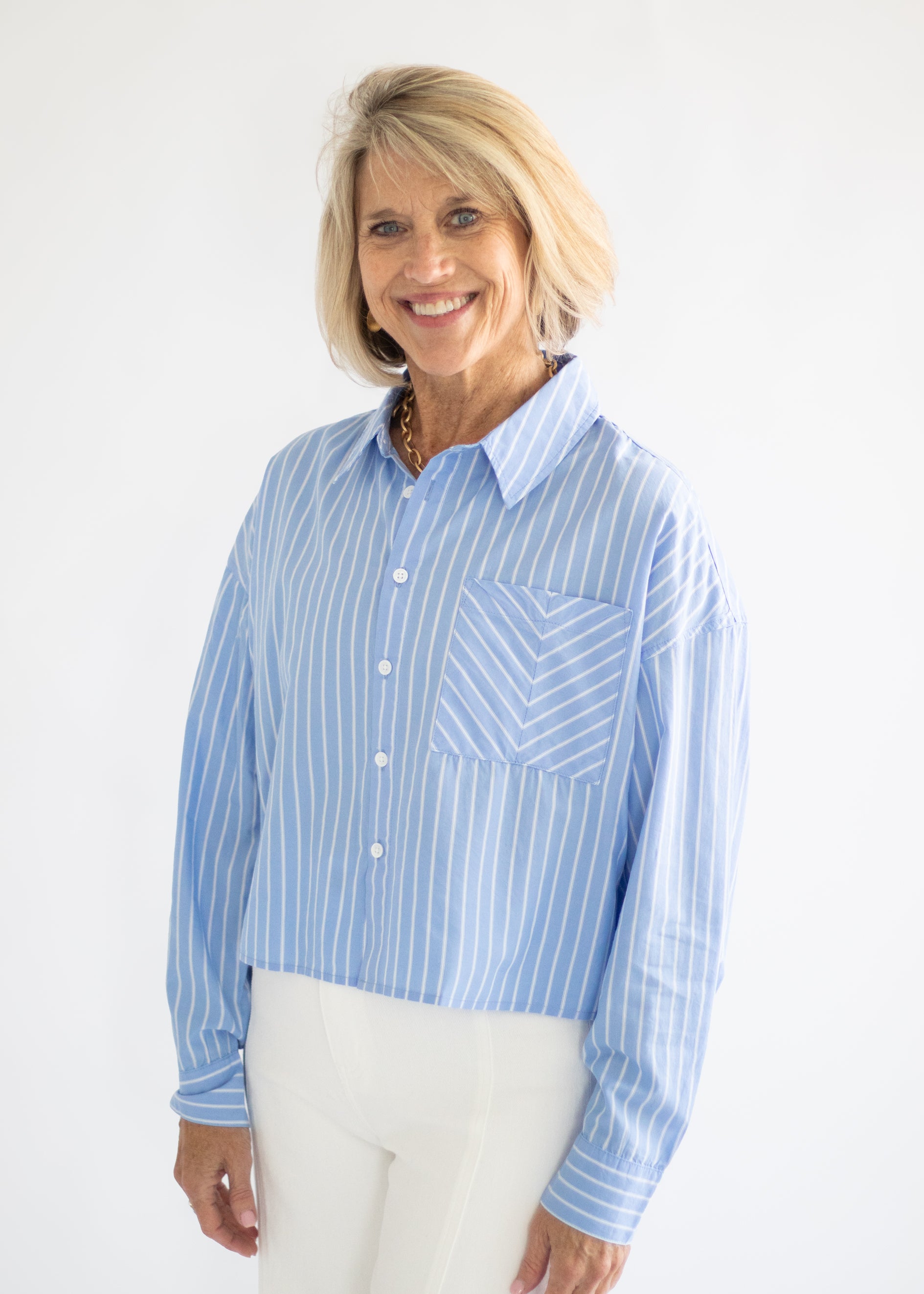 A smiling blonde woman wearing a light blue and white vertically striped button-down shirt with a chest pocket, paired with white pants, against a plain white studio background.